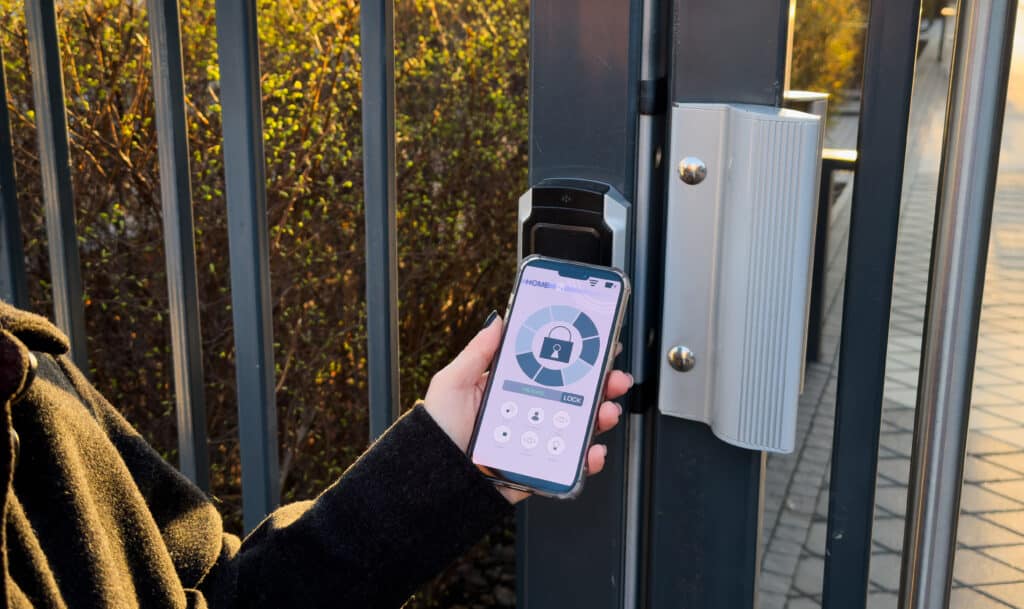 Main d'une personne utilisant un smartphone pour contrôler une serrure connectée sur un portail moderne. L'écran affiche l'application 'HOME' avec un cadenas UNLOCKED et un bouton LOCK.